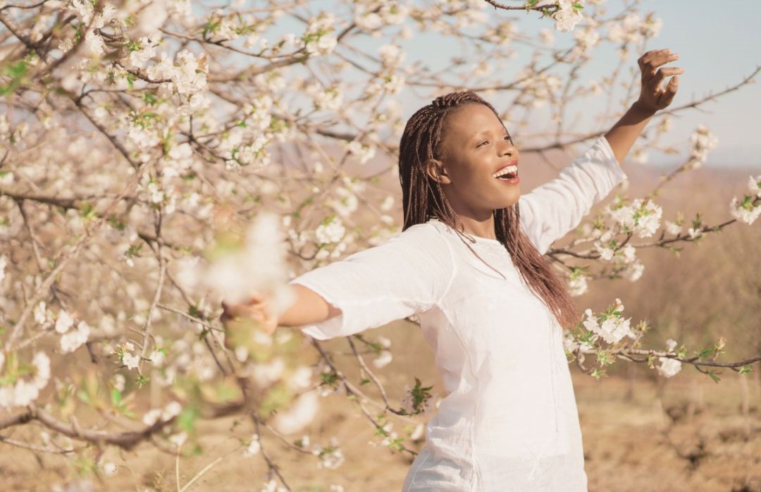 A woman standing with arms outraised and a look of joy on her face in front of a blossom tree