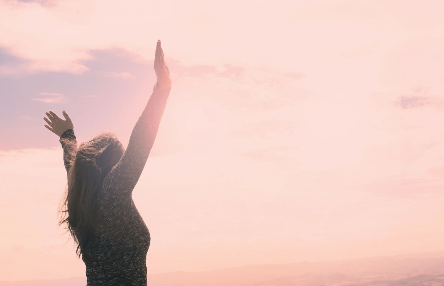 The back of a woman looking out to a pink sky with arms raised looking free and unlocked